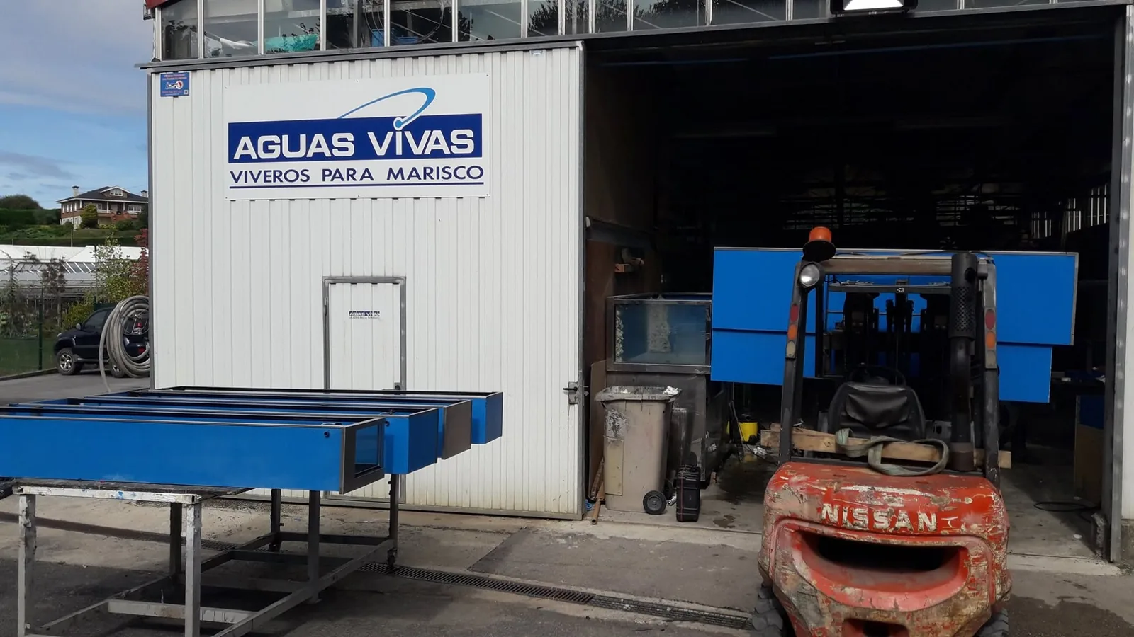 Depósitos azules y una carretilla junto al acceso de la nave de Aguas Vivas