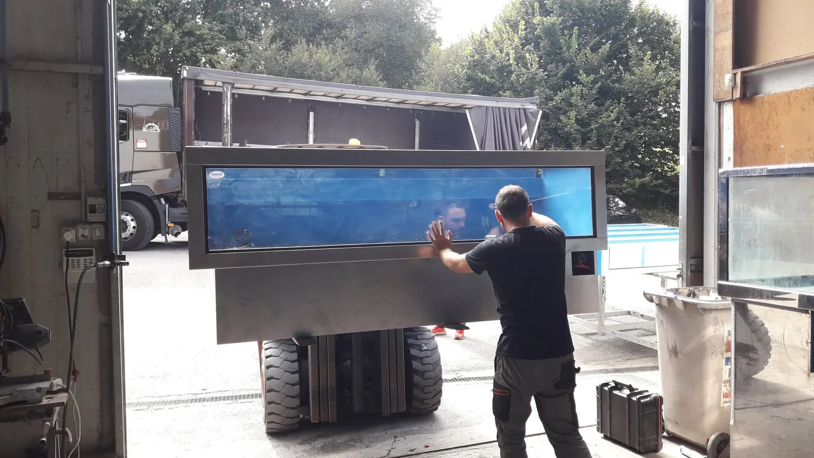 Una persona guía el traslado de un vivero acristalado mediante una carretilla a la entrada del taller